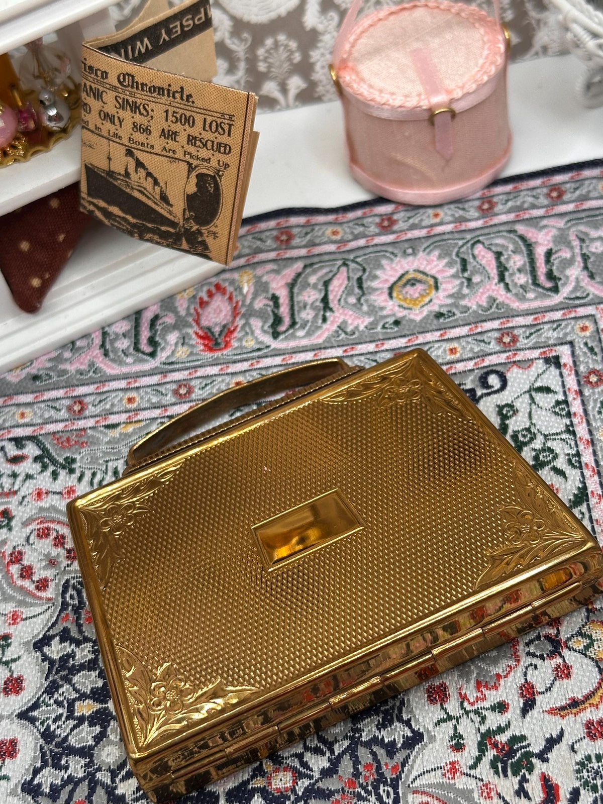 May include: A gold-coloured, rectangular compact with a textured surface and a handle. The compact has floral corner details and a rectangular clasp. A small newspaper clipping with the text "Titanic Sinks" is visible in the background. A pink purse is also in the background.